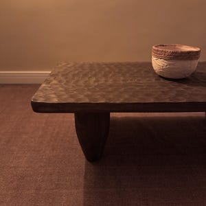 Op de afbeelding: Een donkerbruine houten salontafel met een ruw, getextureerd oppervlak en drie poten. De tafel staat op een bruin tapijt.