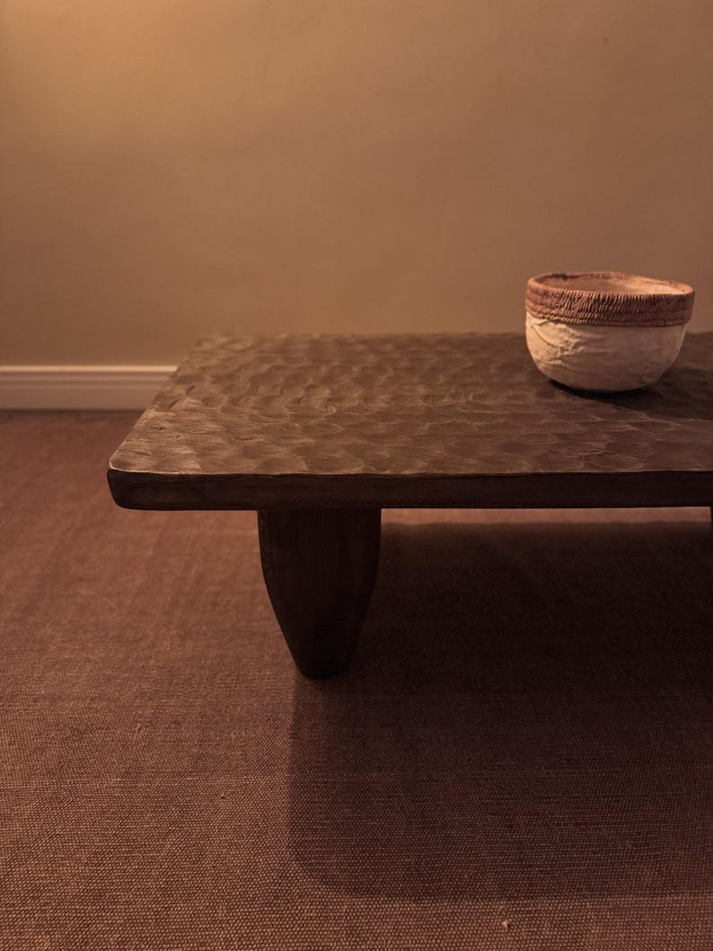 Op de afbeelding: Een donkerbruine houten salontafel met een ruw, getextureerd oppervlak en drie poten. De tafel staat op een bruin tapijt.