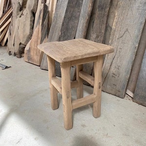 May include: A rustic wooden stool with a square top and four legs. The stool is made of natural wood and has a simple, handcrafted design.
