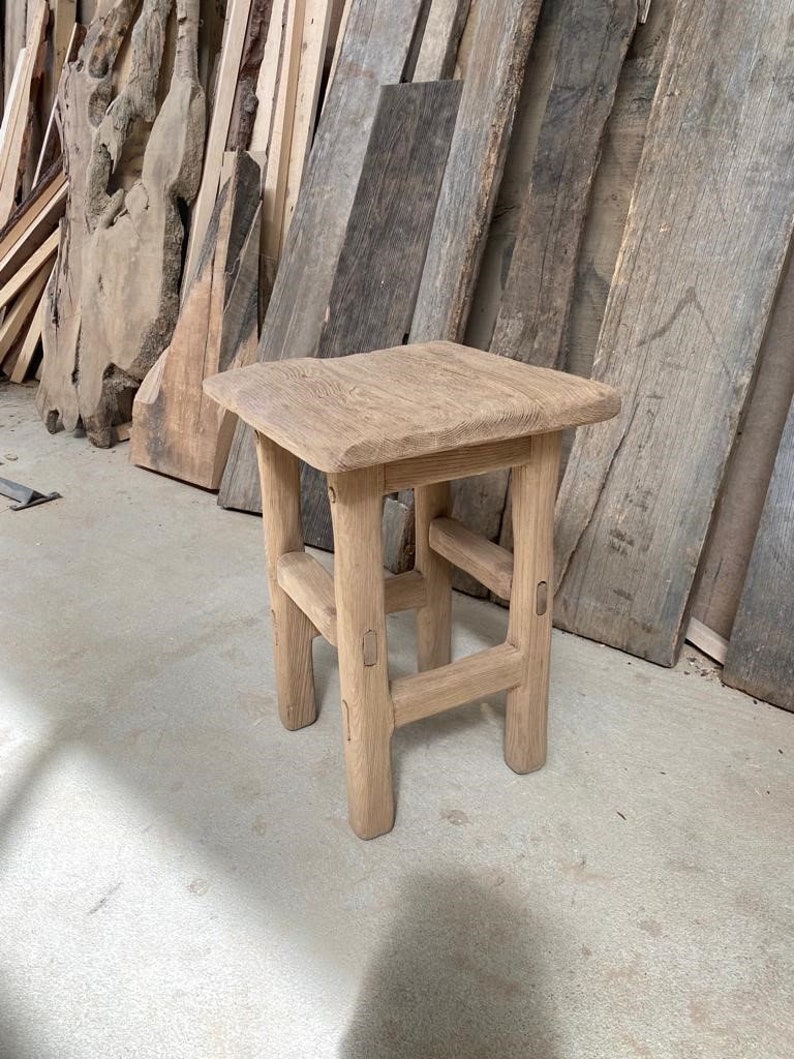 May include: A rustic wooden stool with a square top and four legs. The stool is made of natural wood and has a simple, handcrafted design.