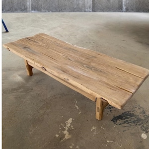 May include: A rectangular, light-colored wooden coffee table with a natural, weathered appearance. The table has four short legs and a slightly uneven surface, showcasing the wood's grain and texture. The table measures approximately 60 inches long.