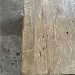 May include: A close-up of a rustic wooden table top with a natural wood grain pattern. The wood is light brown with a weathered finish.