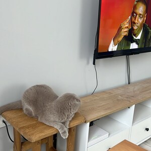 May include: A gray cat sitting on a wooden stool in front of a white entertainment center with a TV playing a movie. The TV is mounted on the wall and has a black frame. The entertainment center has two shelves and a drawer. The cat is looking at the TV.