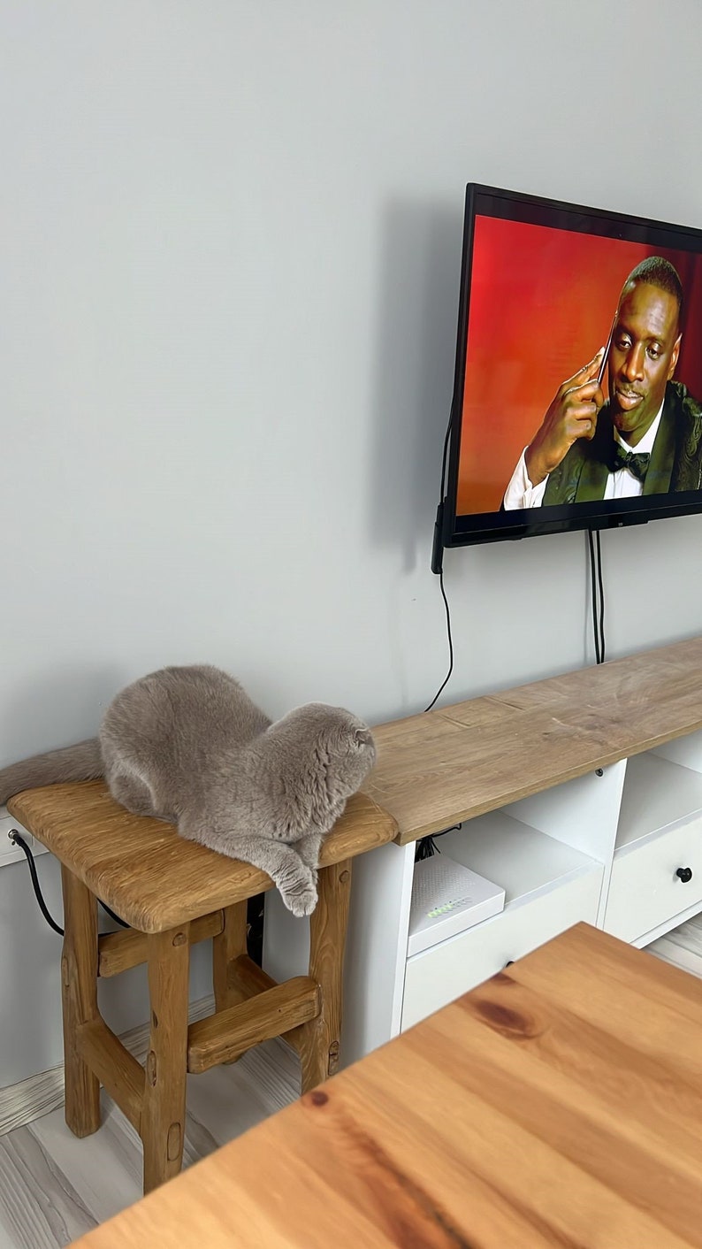 May include: A gray cat sitting on a wooden stool in front of a white entertainment center with a TV playing a movie. The TV is mounted on the wall and has a black frame. The entertainment center has two shelves and a drawer. The cat is looking at the TV.