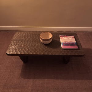 Op de afbeelding: Een donkerbruine houten salontafel met een ruw, getextureerd oppervlak. De tafel heeft vier korte poten en is bedekt met een witte keramische kom en een boek met de titel "Maroc".