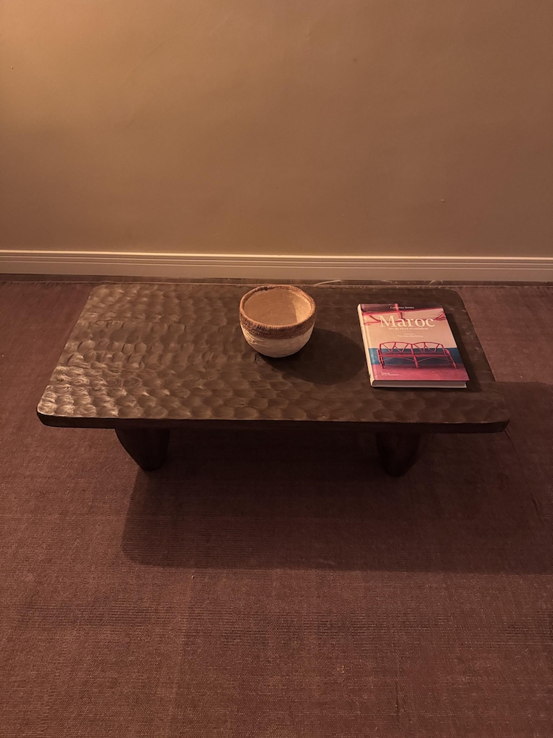 Op de afbeelding: Een donkerbruine houten salontafel met een ruw, getextureerd oppervlak. De tafel heeft vier korte poten en is bedekt met een witte keramische kom en een boek met de titel "Maroc".