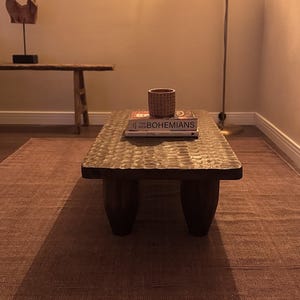 Op de afbeelding: Een houten salontafel met een gegraveerd blad en drie poten. De tafel staat op een bruin tapijt en heeft een boek en een kleine decoratieve pot erop.