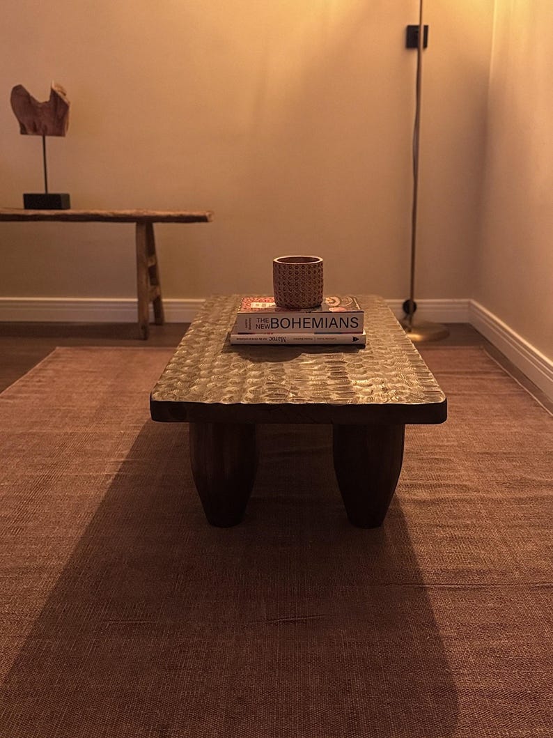 Op de afbeelding: Een houten salontafel met een gegraveerd blad en drie poten. De tafel staat op een bruin tapijt en heeft een boek en een kleine decoratieve pot erop.