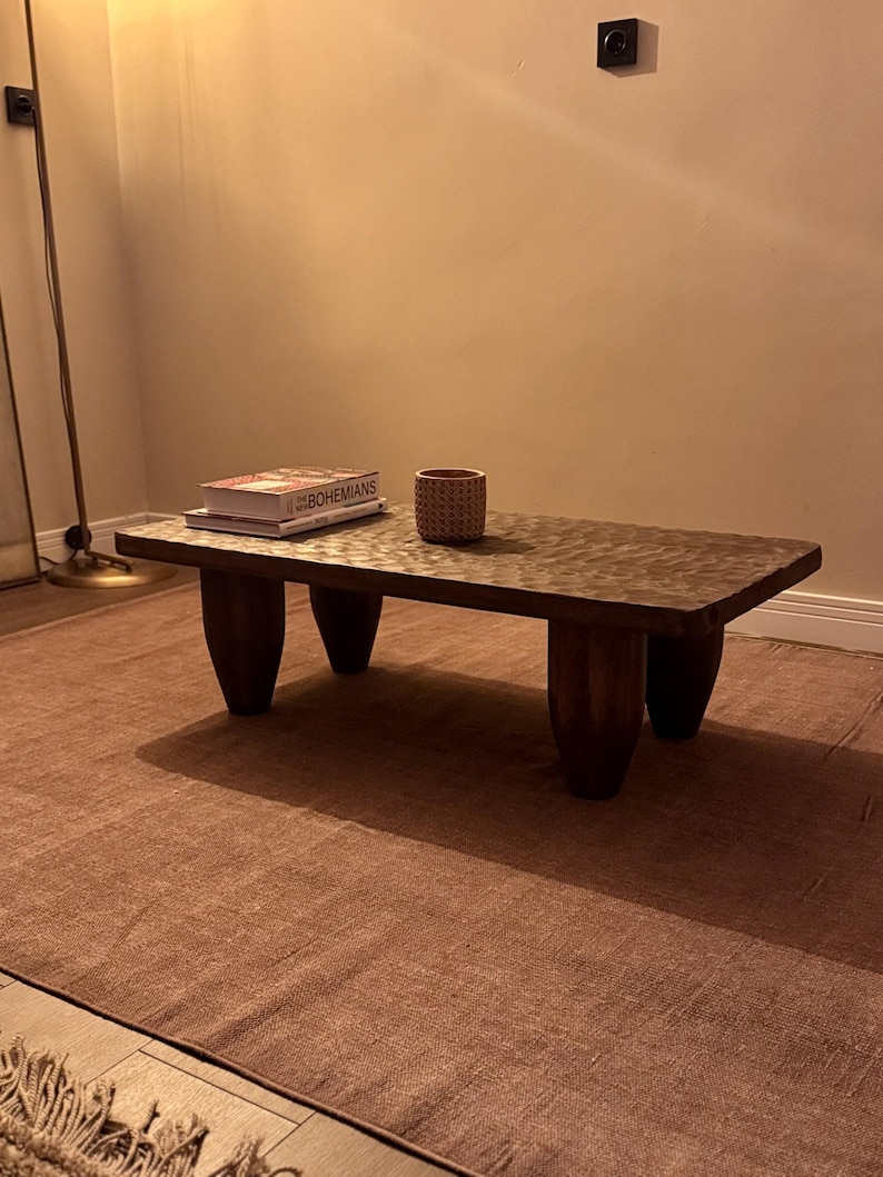 Op de afbeelding: Een houten salontafel met vier dikke, afgeronde poten. De tafel heeft een bruin, getextureerd oppervlak en een boek met de titel "The New Bohemians" erop. Een kleine geweven mand staat naast het boek.