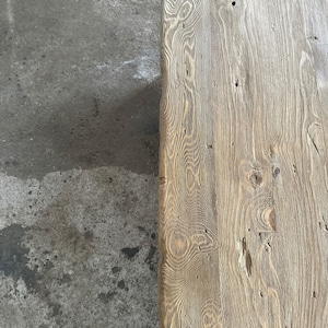 May include: Close-up of a rustic wooden table top with a natural wood grain pattern and knots. The wood is a light brown colour with a weathered finish.