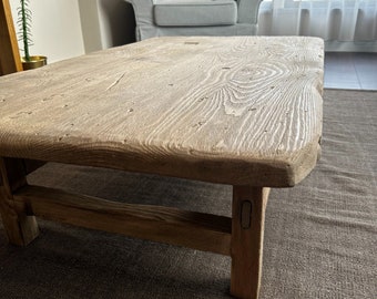 May include: A rustic wooden coffee table with a thick, natural wood top and simple, sturdy legs. The table is sitting on a brown rug.