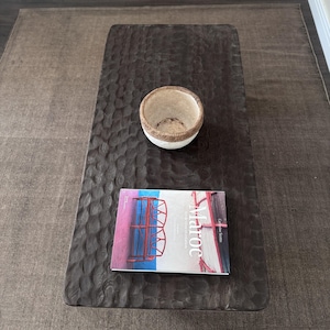 Op de afbeelding: Een donkerbruine houten salontafel met een uniek, getextureerd oppervlak. Een witte kom met een geweven rand staat op de tafel, en een boek getiteld "Maroc" ligt op de tafel. De tafel staat op een bruin tapijt.