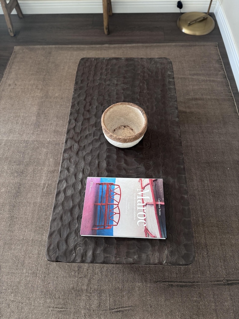 Op de afbeelding: Een donkerbruine houten salontafel met een uniek, getextureerd oppervlak. Een witte kom met een geweven rand staat op de tafel, en een boek getiteld "Maroc" ligt op de tafel. De tafel staat op een bruin tapijt.