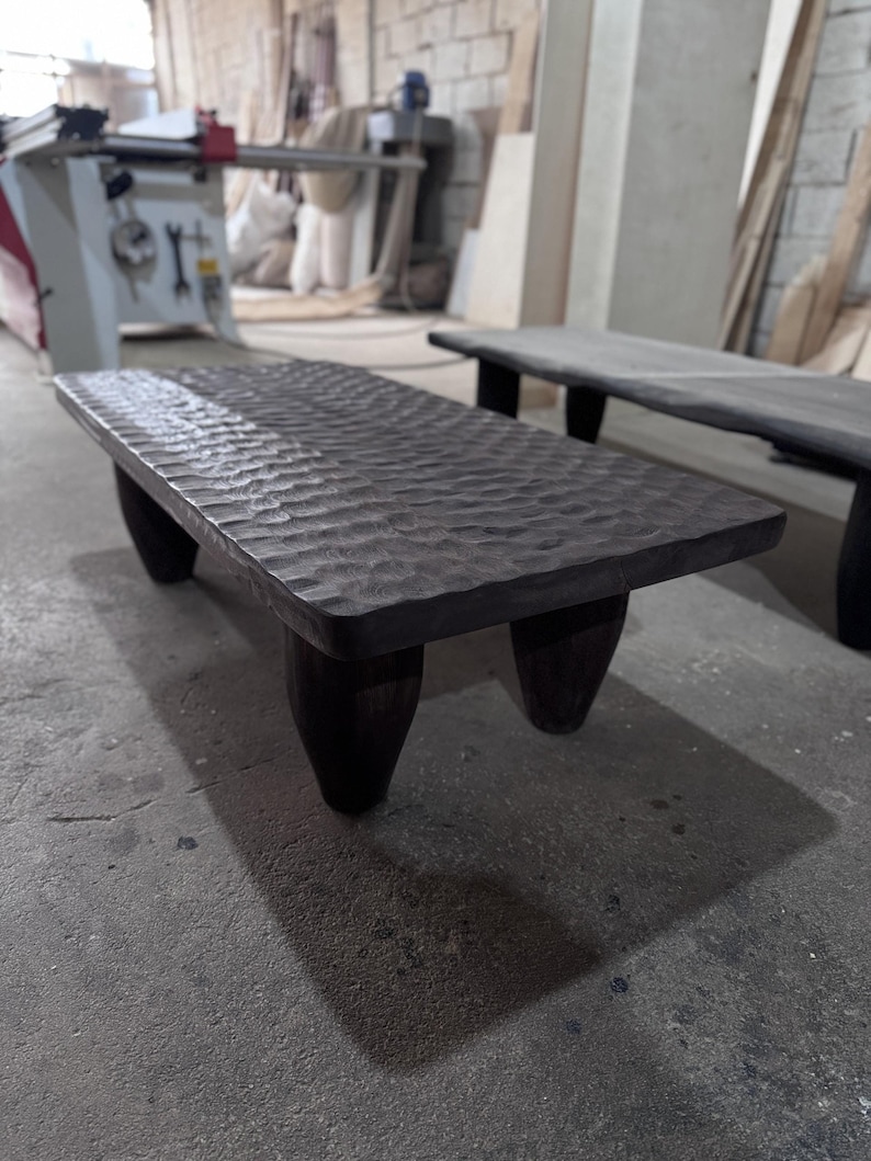 Op de afbeelding: Een donkerbruine houten salontafel met een getextureerd blad en vier poten. De tafel staat in een werkplaats.