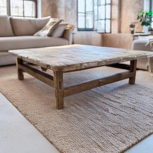 May include: A weathered, square wooden coffee table with a light brown finish. The table is placed on a beige rug in a living room. The table has a simple, sturdy design, with visible wood grain.