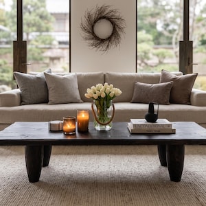May include: A low, dark-stained wooden coffee table with four thick, cylindrical legs, positioned in front of a beige sofa. The table holds a glass vase of white tulips, two lit candles, a stack of books, and a small black decorative object.