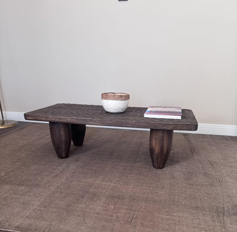 Op de afbeelding: Een donkerbruine houten salontafel met vier dikke, afgeronde poten. De tafel heeft een ruw, getextureerd oppervlak en is versierd met een witte keramische kom en een boek.