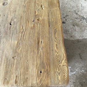 May include: A close-up of a rustic wooden table top with a natural wood grain pattern and knots. The wood has a light brown colour and a rough texture.