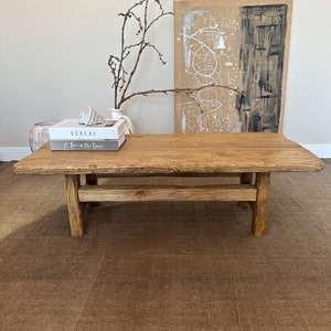 Może przedstawiać: Rustykalny drewniany stolik kawowy z naturalnym wykończeniem drewna. Stolik ma prostokątny blat i jedną p&oacute;łkę pod spodem. Na stoliku leżą dwie książki, jedna zatytułowana "Cereal", a druga "A Tree in the House".
