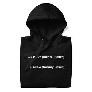 May include: A black hooded sweatshirt featuring white text that reads As above (mental issues) So below (tummy issues). The garment includes a hood with matching black drawstrings and is displayed in a folded position, highlighting the printed text.