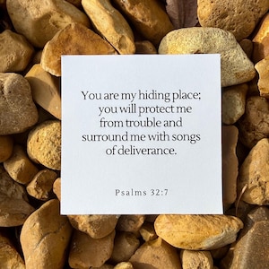 May include: A white card with the text "You are my hiding place; you will protect me from trouble and surround me with songs of deliverance. Psalms 32:7" is placed on a bed of light brown and tan rocks.