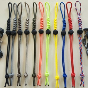 Może przedstawiać: Zestaw 10 kolorowych smyczy do łuku z paracordu z czarnymi koralikami. Smycze są wykonane z paracordu w różnych kolorach i mają węzeł na końcu. Smycze są przymocowane do czarnego spustu łuku.