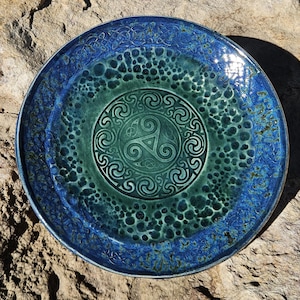 May include: A round ceramic plate featuring a deep blue outer rim with intricate embossed knotwork patterns. The center displays a green glazed surface with a textured, bubbly finish and a central circular medallion containing a triskelion design.