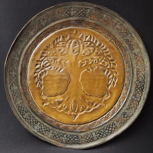 May include: A round decorative plate featuring a central embossed tree design in a warm golden-brown tone. The wide outer rim is finished in a dark, weathered metallic gray with an intricate, repeating Celtic-style knotwork pattern.