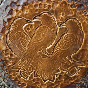 May include: A circular brown leather piece featuring an embossed design of three ravens with intricate knotwork patterns on their bodies. The background has a hammered, textured appearance with dark brown and tan color variations throughout.