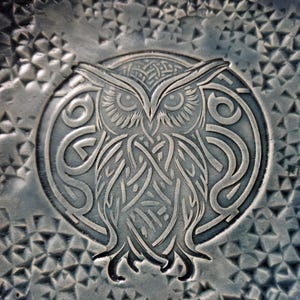 May include: A detailed, close-up image of a circular, embossed design featuring an owl. The owl is the central focus, with intricate Celtic knotwork forming its body and surrounding a circular border. The background has a geometric pattern.