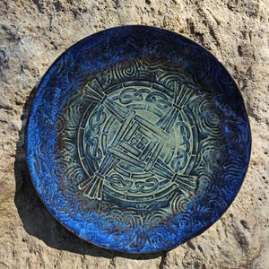 May include: A round ceramic plate featuring a deep blue glazed rim and a central light green medallion. The center displays a raised, embossed geometric cross pattern surrounded by intricate knotwork, with swirling textures throughout the surface.
