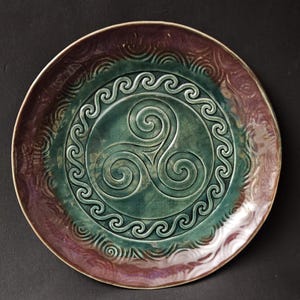 May include: A round ceramic plate featuring a central triskelion symbol embossed in a deep green glaze. The inner circle is bordered by a wave pattern, while the outer rim displays a textured, swirling design in shades of plum and purple.