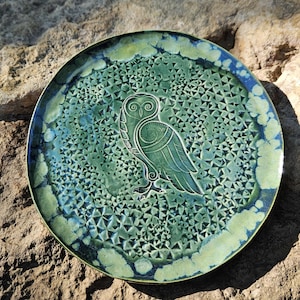 May include: A round ceramic plate featuring a central embossed owl design with intricate spiral details. The surface has a textured, crystalline glaze in shades of deep forest green, lime green, and dark blue, creating a variegated pattern.