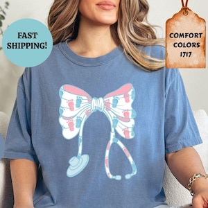 Könnte beinhalten: Ein T-Shirt aus Jeansblau mit einem rosa-blauen Schleifen- und Stethoskop-Design. Die Schleife besteht aus winzigen Fußabdrücken. Der Text "SCHNELLER VERSAND!" befindet sich in einem Kreis über dem Shirt. Der Text "COMFORT COLORS 1717" befindet sich auf einem braunen Etikett.