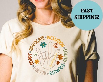 Puede incluir: Una camiseta beige con un gráfico circular que presenta una mano haciendo el signo de la lengua de señas americana para "Te amo". El gráfico también incluye las palabras "Aceptar", "Adaptar", "Abogar", "Igualdad", "Equidad", "Inclusión" y "Fourality" alrededor del signo de la mano.