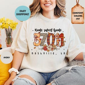 Może przedstawiać: Kremowa koszulka z napisem "Home sweet home" i liczbami "37011" w dekoracyjnym wzorze. Pod spodem znajduje się napis "Nashville, TN". Koszulka to Comfort Colors 1717.
