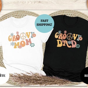 May include: Two t-shirts with a groovy font design. One shirt is white with the text "Groovy Mom" and the other shirt is black with the text "Groovy Dad". Both shirts feature a peace sign and flowers.