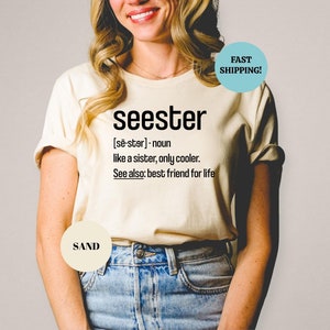 以下が含まれることがあります： 「seester」という言葉の定義が書かれた白いTシャツ。定義は「[sē-ster] - 名詞 姉妹のようなものだが、もっとクール。参考: 一生の親友」