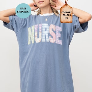 Puede incluir: Una camiseta de gran tamaño de color azul vaquero con la palabra "NURSE" en letras de patchwork coloridas. La camiseta tiene cuello redondo y mangas cortas. La imagen también incluye una etiqueta que dice "Comfort Colors 1717" y un círculo que dice "¡Envío rápido!"