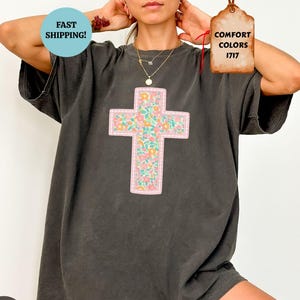 以下が含まれることがあります： ピンクの縁取りと花の十字架のデザインが施された濃いグレーのTシャツ。十字架はカラフルな花柄で埋められています。ゆったりとしたフィット感のシャツで、「Comfort Colors 1717」と表示されています。