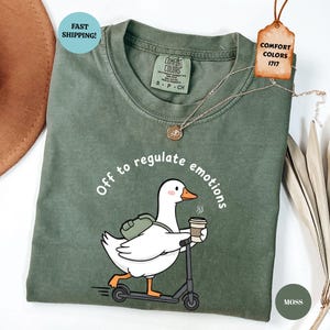 Op de afbeelding: Een mosgroen t-shirt met een cartoon eend op een scooter, met koffie en een rugzak, met de tekst "Off to regulate emotions". Het shirt heeft een ronde hals en korte mouwen. Comfort Colors merk.
