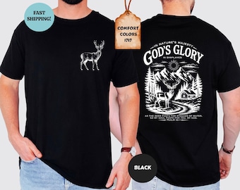 May include: A black t-shirt featuring a white graphic of a deer on the front left chest. The back displays a detailed white illustration of a deer, mountains, trees, and a cabin with the text IN NATURE'S MAJESTY GOD'S GLORY IS DISPLAYED.