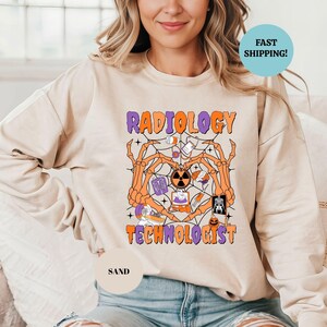 Pode incluir: Camisola cor de areia com as palavras "RADIOLOGY TECHNOLOGIST" em laranja e roxo, com um gráfico temático de Halloween. O gráfico inclui uma teia de aranha, mãos de esqueleto e imagens médicas.