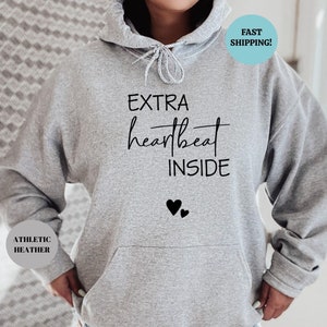Pode incluir: Camisola de moletom cinza com capuz com o texto "EXTRA heartbeat INSIDE" e dois cora&ccedil;&otilde;es pretos abaixo do texto. A camisola &eacute; feita de material heather atl&eacute;tico.