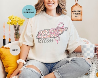 Könnte beinhalten: Ein elfenbeinfarbenes Kurzarm-T-Shirt mit einer Grafik des Bundesstaates Tennessee, die mit einem Blumenmuster in Rosa-, Orange- und Grüntönen gefüllt ist. Eine rosa Schleife ist um den Umriss des Staates gebunden und das Wort Tennessee steht darunter.