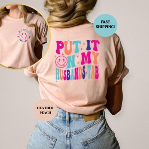 May include: A pink t-shirt with the text "PUT IT ON MY HUSBANDS TAB" in colorful retro letters. The text is surrounded by stars and a smiley face with dollar signs for eyes.