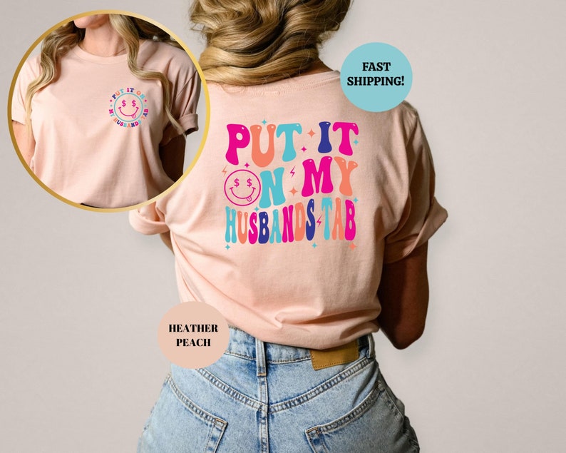 May include: A pink t-shirt with the text "PUT IT ON MY HUSBANDS TAB" in colorful retro letters. The text is surrounded by stars and a smiley face with dollar signs for eyes.