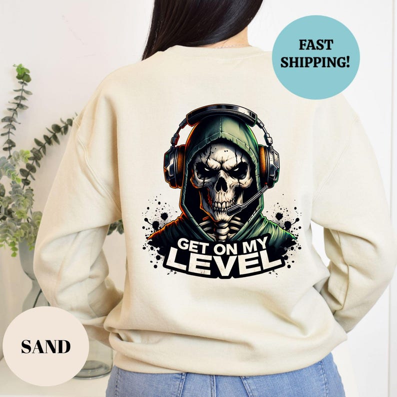 May include: Sand-colored sweatshirt featuring a graphic of a skull wearing a green hoodie and headset. The skull has a microphone and the text "GET ON MY LEVEL" is below. A blue circle with "FAST SHIPPING!" is on the top right.