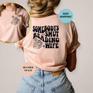 May include: A pink t-shirt with the text "Somebody's Smut Reading Wife" in black, with a skull and stack of books graphic. The shirt is shown on a model wearing blue jeans.