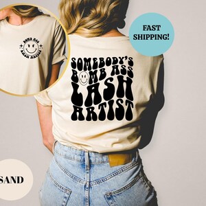 May include: A white t-shirt with a black graphic that says "Somebody's Bomb Ass Lash Artist" in a retro font. The graphic includes a smiley face.